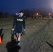 A U.S. Army soldier is performing a kettlebell farmer's carry exercise on an outdoor field during early morning or nighttime hours. The event appears to be a physical fitness training session or assessment, with multiple other soldiers visible in the background performing similar activities. Orange traffic cones mark the course lanes on a grass and dirt field illuminated by artificial lighting.