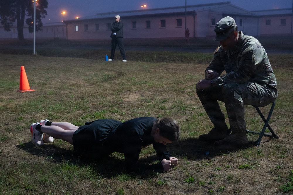 A military fitness assessment is taking place outdoors on a grassy field during early morning or dusk in foggy conditions. A person in civilian athletic wear is performing a prone plank or push-up exercise while a soldier in camouflage uniform observes and evaluates from a small folding stool. A third individual stands in the background near military-style buildings.