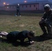 A military fitness assessment is taking place outdoors on a grassy field during early morning or dusk in foggy conditions. A person in civilian athletic wear is performing a prone plank or push-up exercise while a soldier in camouflage uniform observes and evaluates from a small folding stool. A third individual stands in the background near military-style buildings.