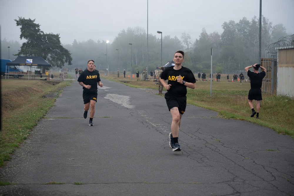Two U.S. Army soldiers in black PT (Physical Training) uniforms are running on a paved path during what appears to be a physical fitness test or training event. The setting is a military installation on an overcast, foggy day with other personnel visible in the background. A blue tent with signage and additional soldiers can be seen further down the course.