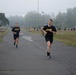 Two U.S. Army soldiers in black PT (Physical Training) uniforms are running on a paved path during what appears to be a physical fitness test or training event. The setting is a military installation on an overcast, foggy day with other personnel visible in the background. A blue tent with signage and additional soldiers can be seen further down the course.