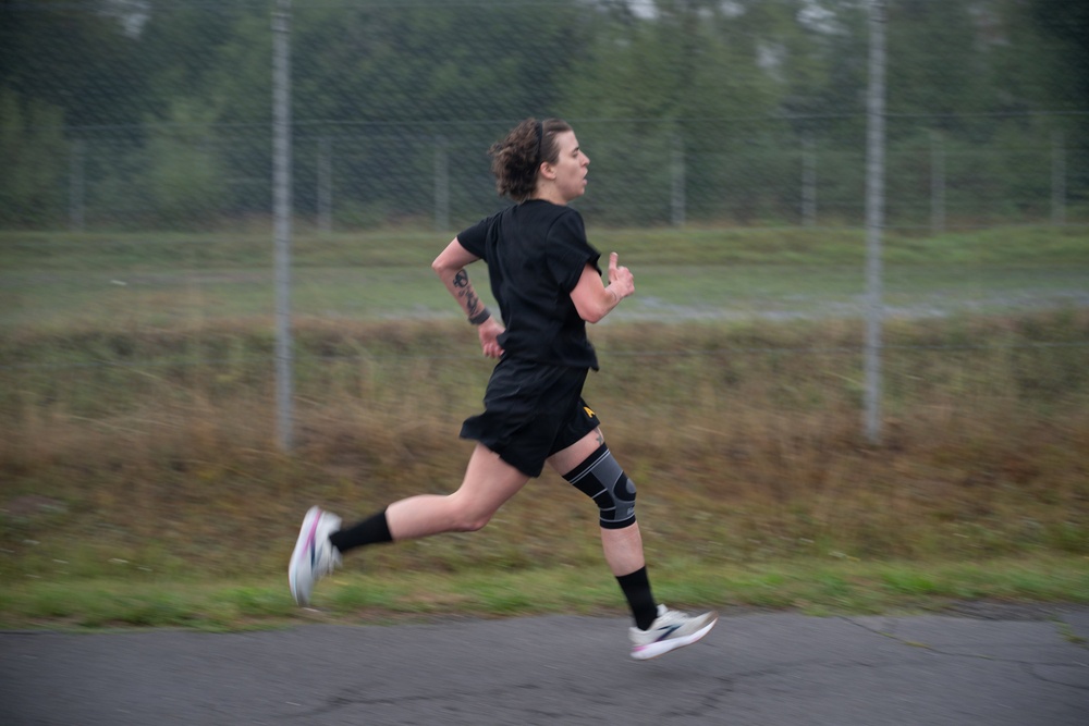 A person is running outdoors on a paved surface, wearing all-black athletic clothing including a t-shirt and shorts. They have a knee brace on their right knee and black compression socks, along with white running shoes. The background shows a chain-link fence, overgrown grass, and trees in an overcast setting.