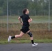 A person is running outdoors on a paved surface, wearing all-black athletic clothing including a t-shirt and shorts. They have a knee brace on their right knee and black compression socks, along with white running shoes. The background shows a chain-link fence, overgrown grass, and trees in an overcast setting.