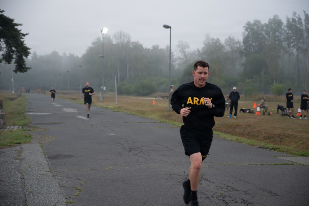 A group of U.S. Army soldiers are conducting a physical fitness test or training run on a paved road at what appears to be a military installation. The foreground shows a male soldier in standard Army PT (Physical Training) uniform running toward the camera, with several others visible behind him. The setting is overcast and foggy, suggesting an early morning or cool-weather training session.