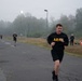 A group of U.S. Army soldiers are conducting a physical fitness test or training run on a paved road at what appears to be a military installation. The foreground shows a male soldier in standard Army PT (Physical Training) uniform running toward the camera, with several others visible behind him. The setting is overcast and foggy, suggesting an early morning or cool-weather training session.
