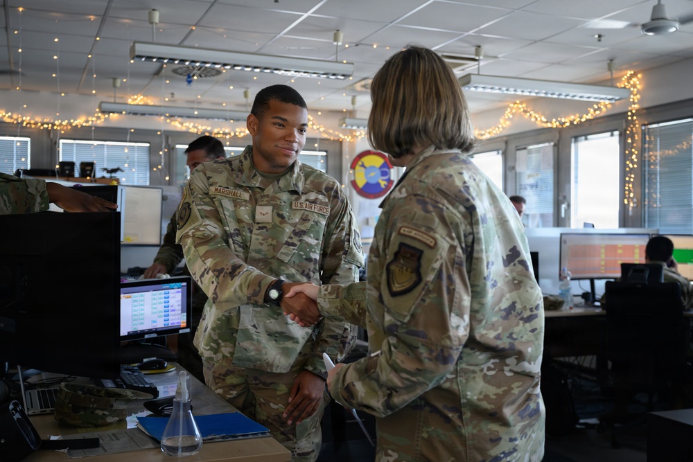 Two U.S. Air Force personnel in OCP (Operational Camouflage Pattern) uniforms are shaking hands in what appears to be a military operations or administrative office. The airman facing the camera has a name tape reading 'MARSHALL' and 'U.S. AIR FORCE' tape visible. The office environment is decorated with string lights, suggesting a holiday period, and contains multiple workstations with computer monitors.