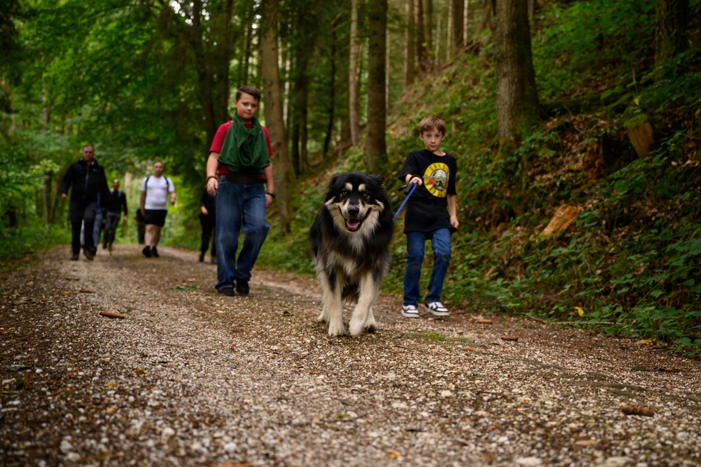 A group of people are hiking along a gravel forest trail surrounded by dense green trees. A young boy in a black Guns N' Roses t-shirt is leading a large black-and-white fluffy dog on a blue leash. Several other individuals, including another youth and two adults, follow behind on the same path.