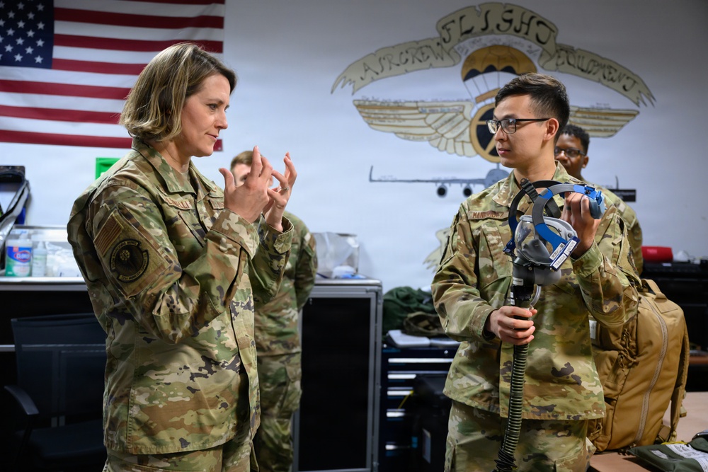 Two U.S. Air Force personnel in OCP (Operational Camouflage Pattern) uniforms are engaged in a discussion inside what appears to be an Aircrew Flight Equipment (AFE) shop. The service member on the right is holding an oxygen mask/breathing apparatus while the one on the left appears to be explaining or instructing. A U.S. flag and an Aircrew Flight Equipment unit emblem are visible on the wall behind them.