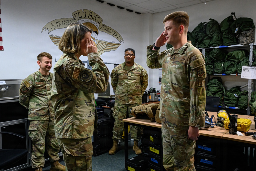 Four military personnel in OCP (Operational Camouflage Pattern) uniforms are gathered in what appears to be an Aircraft Flight Equipment room, as indicated by the unit insignia on the wall. Two individuals in the foreground are exchanging salutes, suggesting a formal military ceremony or greeting. Two other service members stand in the background observing the interaction.