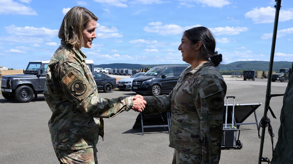Two female military personnel in OCP (Operational Camouflage Pattern) uniforms are shaking hands on what appears to be a military airfield or tarmac. The individual on the left displays a circular patch on her left shoulder, suggesting a higher command or special unit affiliation. The setting appears to be an outdoor military installation with vehicles and rolling hills visible in the background.