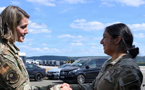 Two female military personnel in OCP (Operational Camouflage Pattern) uniforms are shaking hands on what appears to be a military airfield or tarmac. The individual on the left displays a circular patch on her left shoulder, suggesting a higher command or special unit affiliation. The setting appears to be an outdoor military installation with vehicles and rolling hills visible in the background.