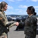 Two female military personnel in OCP (Operational Camouflage Pattern) uniforms are shaking hands on what appears to be a military airfield or tarmac. The individual on the left displays a circular patch on her left shoulder, suggesting a higher command or special unit affiliation. The setting appears to be an outdoor military installation with vehicles and rolling hills visible in the background.