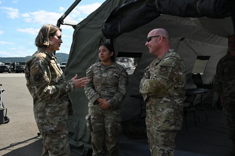 Three U.S. military personnel in OCP (Operational Camouflage Pattern) uniforms are engaged in conversation near a military field tent. The scene appears to be an outdoor military installation or exercise area, with vehicles and equipment visible in the background. A senior female service member on the left appears to be briefing or speaking with two other personnel.