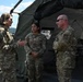 Three U.S. military personnel in OCP (Operational Camouflage Pattern) uniforms are engaged in conversation near a military field tent. The scene appears to be an outdoor military installation or exercise area, with vehicles and equipment visible in the background. A senior female service member on the left appears to be briefing or speaking with two other personnel.