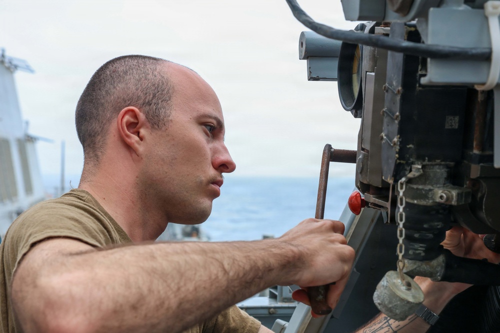 A young male service member in a olive/tan t-shirt is performing maintenance or inspection work on a piece of naval deck equipment. The work is taking place aboard a military vessel at sea, with ocean and overcast sky visible in the background. The individual is focused intently on the mechanical component he is handling.