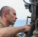 A young male service member in a olive/tan t-shirt is performing maintenance or inspection work on a piece of naval deck equipment. The work is taking place aboard a military vessel at sea, with ocean and overcast sky visible in the background. The individual is focused intently on the mechanical component he is handling.