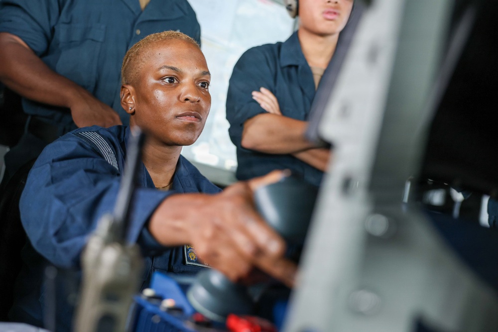 A woman in dark blue military or naval coveralls is operating controls, likely at a helm or equipment station, while two other personnel in similar uniforms observe from behind. The primary subject is focused intently on the equipment in front of her. The setting appears to be aboard a vessel or in a technical operational environment.