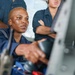 A woman in dark blue military or naval coveralls is operating controls, likely at a helm or equipment station, while two other personnel in similar uniforms observe from behind. The primary subject is focused intently on the equipment in front of her. The setting appears to be aboard a vessel or in a technical operational environment.