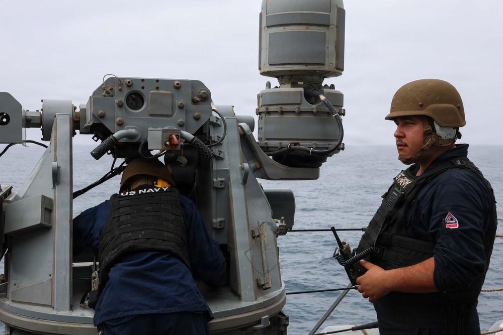 Two U.S. Navy personnel are stationed on the deck of a naval vessel at sea, positioned near a weapon or sensor system mount. One sailor is wearing a vest labeled 'US NAVY' and yellow ear protection, while the other wears a tan combat helmet and tactical vest and holds a radio. The overcast sky and open ocean are visible in the background.