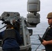 Two U.S. Navy personnel are stationed on the deck of a naval vessel at sea, positioned near a weapon or sensor system mount. One sailor is wearing a vest labeled 'US NAVY' and yellow ear protection, while the other wears a tan combat helmet and tactical vest and holds a radio. The overcast sky and open ocean are visible in the background.