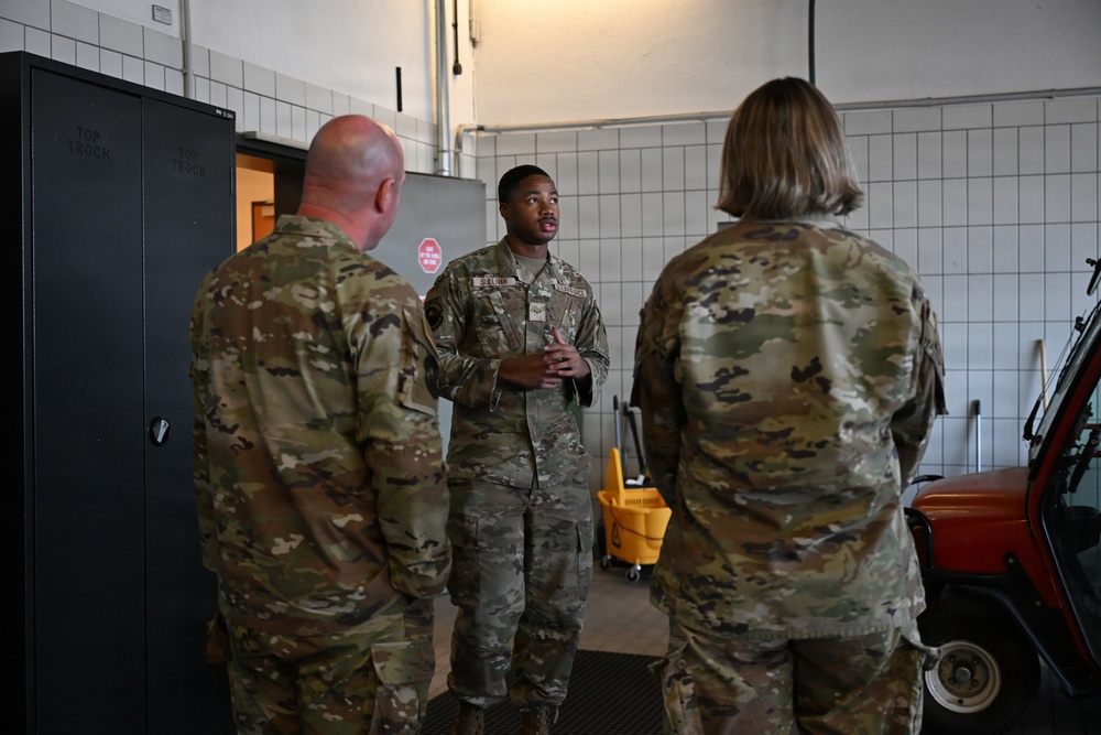 Three military personnel in OCP (Operational Camouflage Pattern) uniforms are engaged in a conversation inside what appears to be a maintenance or utility facility. The central figure, facing the camera, appears to be briefing or explaining something to the other two individuals whose backs are turned. The setting suggests an indoor military installation, possibly a vehicle maintenance bay or storage area.