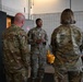 Three military personnel in OCP (Operational Camouflage Pattern) uniforms are engaged in a conversation inside what appears to be a maintenance or utility facility. The central figure, facing the camera, appears to be briefing or explaining something to the other two individuals whose backs are turned. The setting suggests an indoor military installation, possibly a vehicle maintenance bay or storage area.