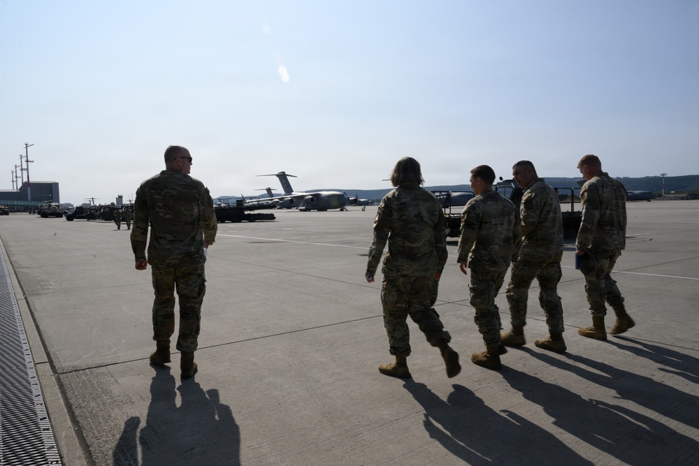 Five military personnel in camouflage uniforms are walking across a large airfield tarmac toward parked military aircraft in the background. The scene appears to be at a military air base with large transport aircraft, likely C-17 Globemasters, visible on the flight line. Equipment and vehicles are staged along the left side of the tarmac.