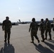Five military personnel in camouflage uniforms are walking across a large airfield tarmac toward parked military aircraft in the background. The scene appears to be at a military air base with large transport aircraft, likely C-17 Globemasters, visible on the flight line. Equipment and vehicles are staged along the left side of the tarmac.