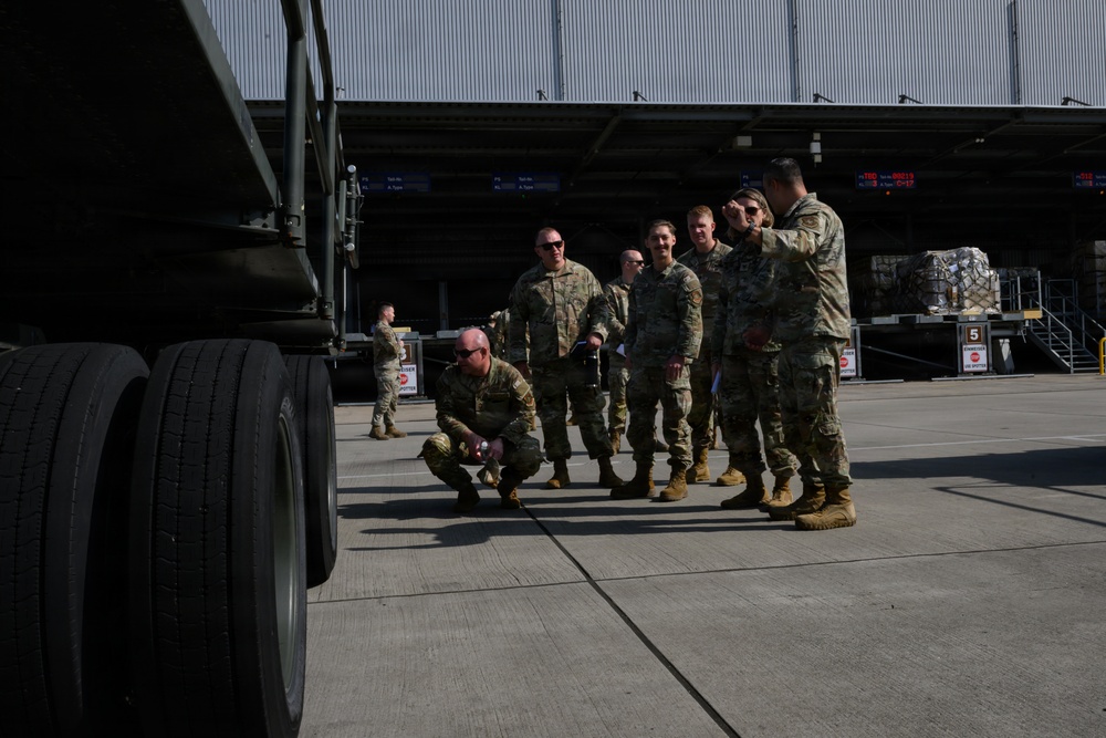 A group of military personnel in camouflage uniforms are gathered near a large military truck on what appears to be a logistics or cargo facility apron. One soldier is crouching near the vehicle's tires while others stand in a group, apparently receiving a briefing or conducting an inspection. The background shows a large warehouse or hangar with cargo pallets and loading equipment visible.