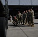 A group of military personnel in camouflage uniforms are gathered near a large military truck on what appears to be a logistics or cargo facility apron. One soldier is crouching near the vehicle's tires while others stand in a group, apparently receiving a briefing or conducting an inspection. The background shows a large warehouse or hangar with cargo pallets and loading equipment visible.