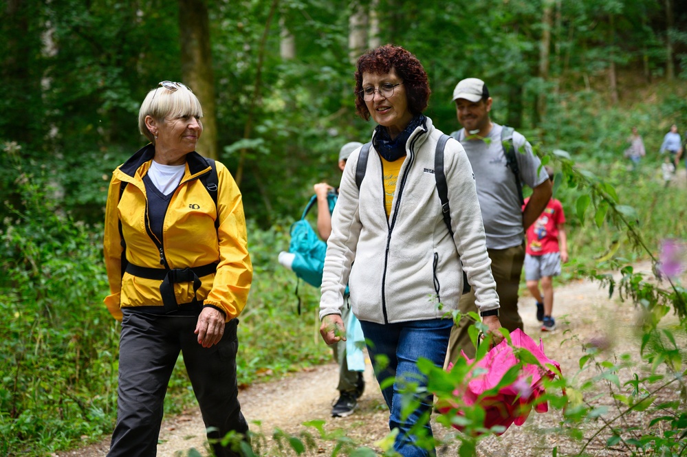A group of people are hiking along a forest trail surrounded by lush green vegetation. Two women are prominently featured in the foreground, appearing to be engaged in conversation while walking. Additional hikers are visible in the background on the same trail.