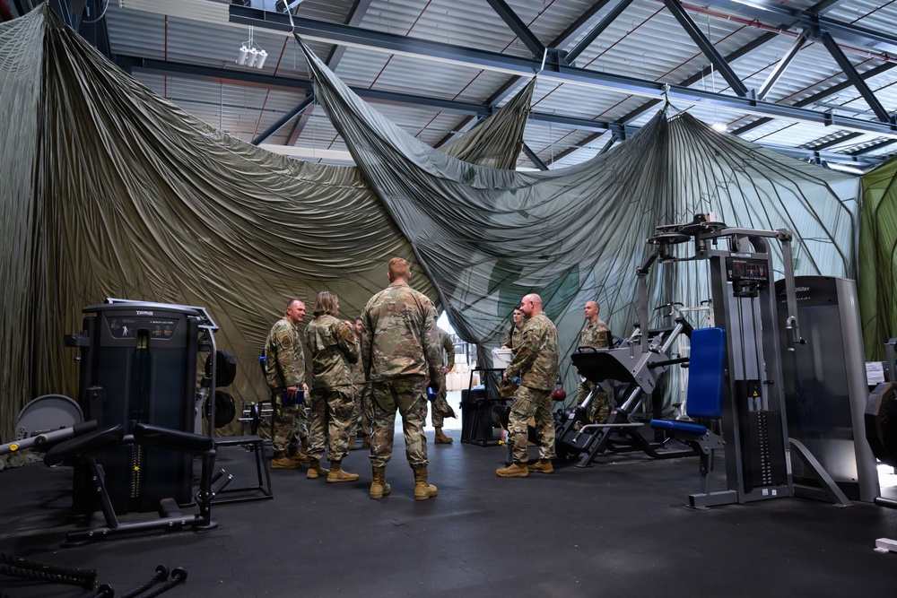 A group of military personnel in camouflage uniforms are gathered inside a large industrial or warehouse-style building that has been converted into a makeshift fitness facility. Large olive-green military canvas or netting is draped from the ceiling, partially dividing the space. Various gym equipment including weight machines and exercise apparatus are visible throughout the area.