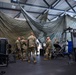 A group of military personnel in camouflage uniforms are gathered inside a large industrial or warehouse-style building that has been converted into a makeshift fitness facility. Large olive-green military canvas or netting is draped from the ceiling, partially dividing the space. Various gym equipment including weight machines and exercise apparatus are visible throughout the area.