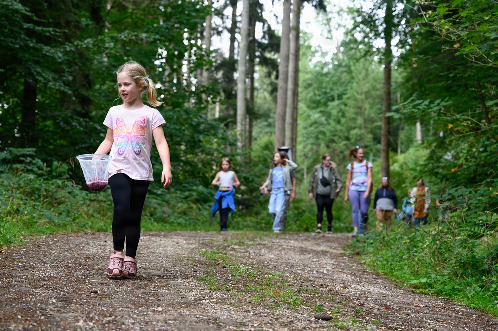 A group of children and adults are walking along a forest trail, likely on a nature hike or outdoor excursion. A young blonde girl in the foreground leads the group, carrying a clear plastic cup containing what appears to be dark berries. The lush green forest setting and casual attire suggest a recreational outdoor activity during summer or early autumn.
