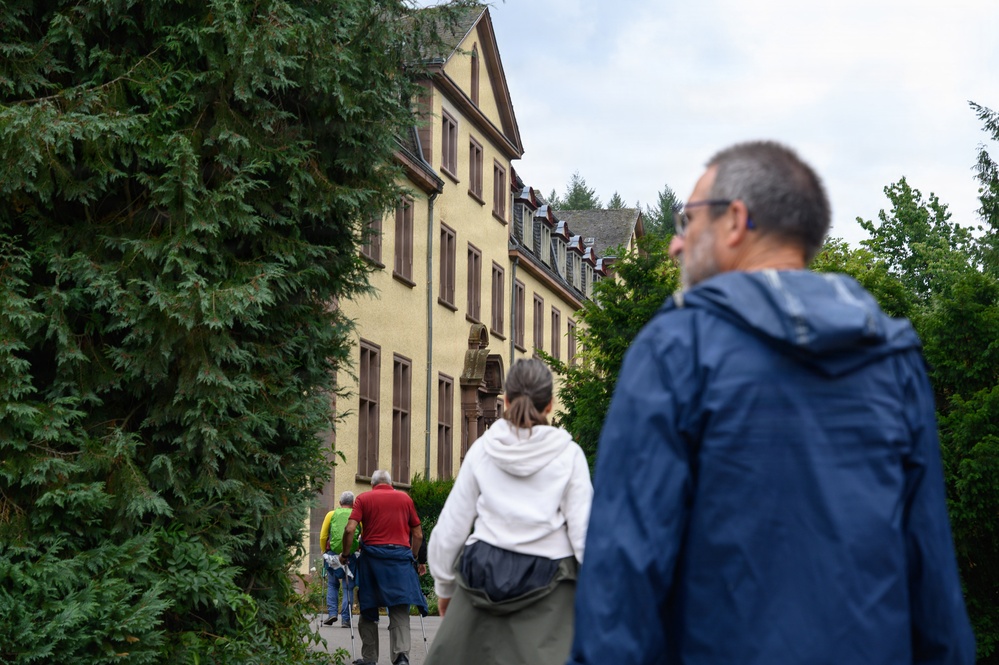 A group of people are walking toward a large, multi-story yellow building with brown-trimmed windows and a mansard-style roof, surrounded by dense green trees. The scene appears to be an outdoor guided tour or group visit to what looks like a historic European-style institutional or residential building. The foreground subjects are out of focus, drawing attention to the building in the background.