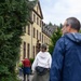 A group of people are walking toward a large, multi-story yellow building with brown-trimmed windows and a mansard-style roof, surrounded by dense green trees. The scene appears to be an outdoor guided tour or group visit to what looks like a historic European-style institutional or residential building. The foreground subjects are out of focus, drawing attention to the building in the background.