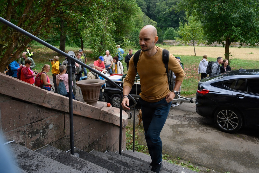 A bald man wearing a mustard yellow sweater and dark jeans is ascending stone steps while carrying a backpack. Behind him, a crowd of people is gathered in an outdoor area near trees and a grassy field, suggesting a group event or tour. A dark-colored BMW SUV is parked nearby on a paved surface.