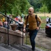 A bald man wearing a mustard yellow sweater and dark jeans is ascending stone steps while carrying a backpack. Behind him, a crowd of people is gathered in an outdoor area near trees and a grassy field, suggesting a group event or tour. A dark-colored BMW SUV is parked nearby on a paved surface.