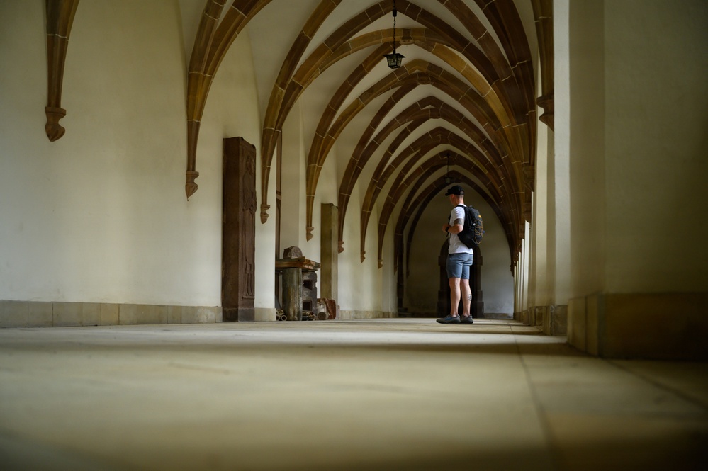 A lone visitor stands at the far end of a Gothic vaulted corridor, likely inside a medieval monastery, cathedral cloister, or historic castle. The hallway features repeating ribbed stone arches that create a strong perspective vanishing point toward the figure. The space is well-lit with natural and artificial light, highlighting the architectural details of the cream-colored plaster walls and sandstone ribs.