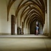 A lone visitor stands at the far end of a Gothic vaulted corridor, likely inside a medieval monastery, cathedral cloister, or historic castle. The hallway features repeating ribbed stone arches that create a strong perspective vanishing point toward the figure. The space is well-lit with natural and artificial light, highlighting the architectural details of the cream-colored plaster walls and sandstone ribs.