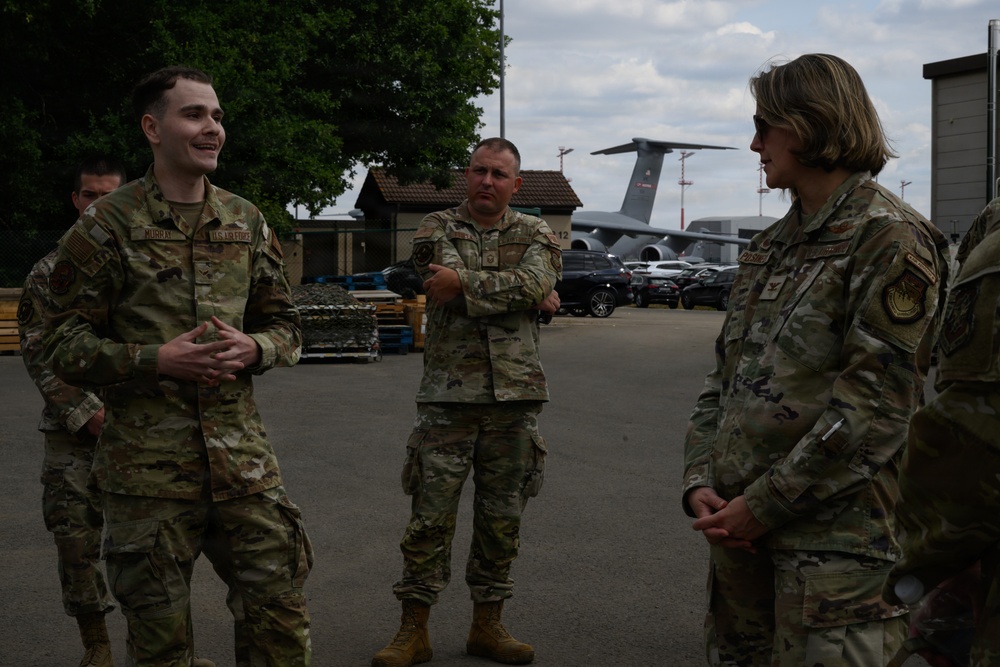 Three U.S. Air Force personnel in OCP (Operational Camouflage Pattern) uniforms are engaged in conversation outdoors on what appears to be a military airfield or base. A younger airman on the left appears to be briefing or speaking to a senior female officer on the right, while a third service member stands in the background with arms crossed. Military aircraft, including what appears to be a C-17 or similar large transport aircraft, are visible in the background.