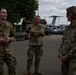 Three U.S. Air Force personnel in OCP (Operational Camouflage Pattern) uniforms are engaged in conversation outdoors on what appears to be a military airfield or base. A younger airman on the left appears to be briefing or speaking to a senior female officer on the right, while a third service member stands in the background with arms crossed. Military aircraft, including what appears to be a C-17 or similar large transport aircraft, are visible in the background.