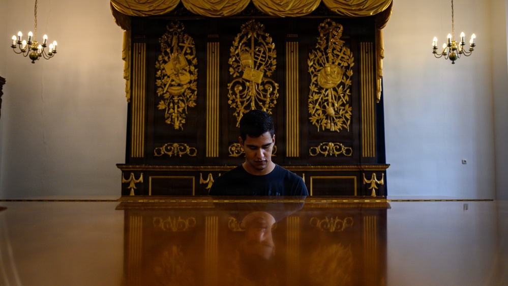 A young man sits at a highly polished grand piano in an ornate, formal room. Behind him is an elaborate dark wood cabinet with intricate gold gilded decorations featuring musical instruments and baroque motifs. The room is flanked by two candelabra-style chandeliers, suggesting a historic palace or concert hall setting.