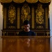 A young man sits at a highly polished grand piano in an ornate, formal room. Behind him is an elaborate dark wood cabinet with intricate gold gilded decorations featuring musical instruments and baroque motifs. The room is flanked by two candelabra-style chandeliers, suggesting a historic palace or concert hall setting.
