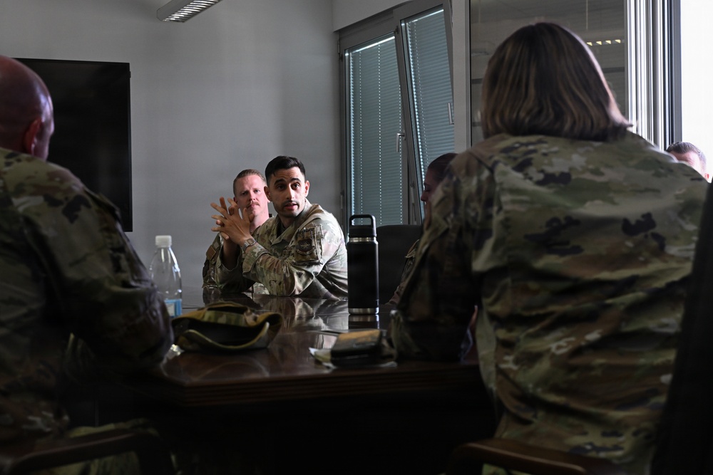 A group of military personnel in camouflage uniforms are seated around a conference table engaged in a discussion or briefing. One individual in the center appears to be actively speaking or gesturing with their hands. The setting appears to be an indoor meeting room with a window and what appears to be a dark display screen in the background.