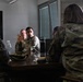 A group of military personnel in camouflage uniforms are seated around a conference table engaged in a discussion or briefing. One individual in the center appears to be actively speaking or gesturing with their hands. The setting appears to be an indoor meeting room with a window and what appears to be a dark display screen in the background.