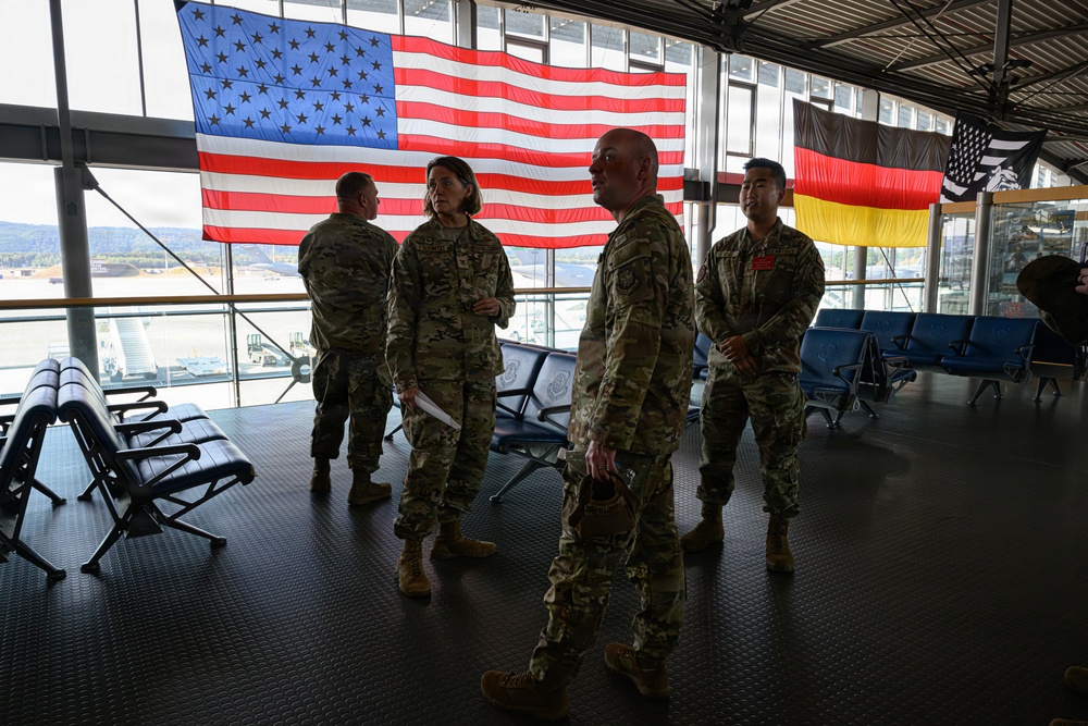 Several U.S. military personnel in OCP (Operational Camouflage Pattern) uniforms are gathered in what appears to be a military terminal or airport lounge. A large American flag is prominently displayed on the back wall, alongside a German flag and what appears to be a unit or organizational flag. The setting suggests a military installation in Germany, likely Ramstein Air Base or a similar U.S. facility.