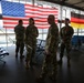 Several U.S. military personnel in OCP (Operational Camouflage Pattern) uniforms are gathered in what appears to be a military terminal or airport lounge. A large American flag is prominently displayed on the back wall, alongside a German flag and what appears to be a unit or organizational flag. The setting suggests a military installation in Germany, likely Ramstein Air Base or a similar U.S. facility.