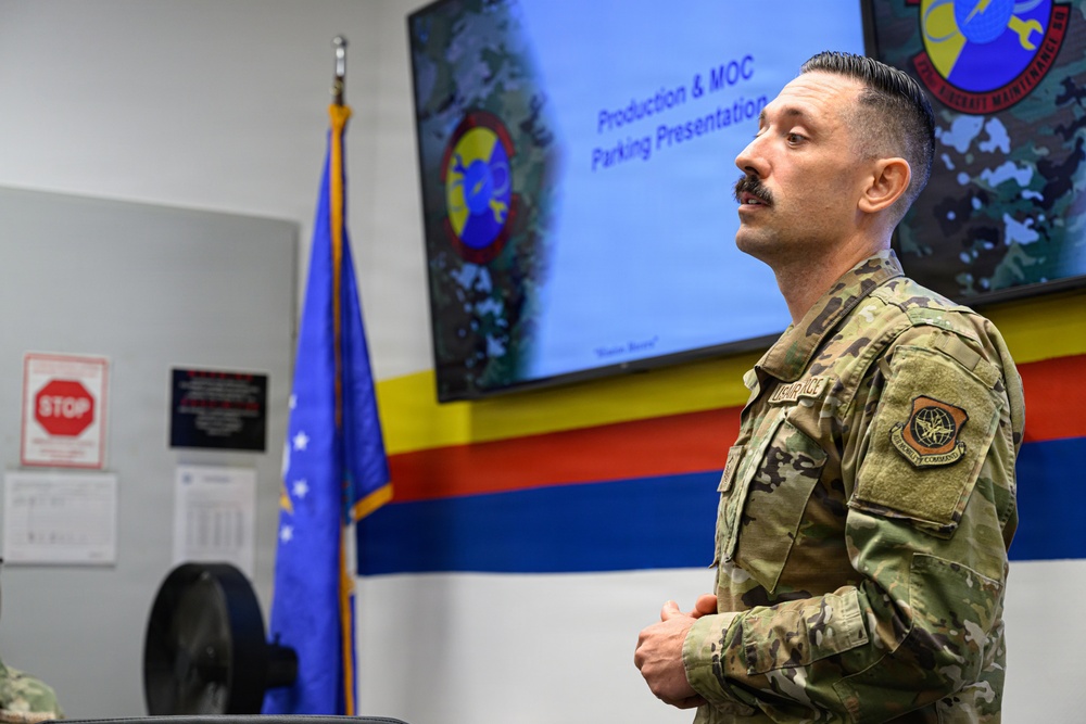 A U.S. Air Force service member in OCP (Operational Camouflage Pattern) uniform stands in a briefing room, apparently presenting or listening to a 'Production &amp; MOC Parking Presentation' displayed on a large screen behind him. The setting appears to be a military operations or maintenance facility, indicated by unit insignia, colored wall stripes, and a U.S. Air Force flag. The individual has a mustache and short dark hair, with arms crossed in an attentive posture.