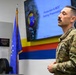 A U.S. Air Force service member in OCP (Operational Camouflage Pattern) uniform stands in a briefing room, apparently presenting or listening to a 'Production &amp; MOC Parking Presentation' displayed on a large screen behind him. The setting appears to be a military operations or maintenance facility, indicated by unit insignia, colored wall stripes, and a U.S. Air Force flag. The individual has a mustache and short dark hair, with arms crossed in an attentive posture.